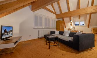 A modern living room with wooden flooring and high ceiling beams. There is a comfortable sofa, a coffee table, and a television.