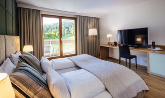 A modern hotel room with a double bed, a desk, and a television. Large windows offer a view of the nature outside.