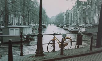 A yellow bicycle is standing at a railing next to a canal. In the background, gray buildings and boats can be seen.