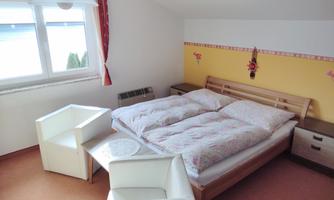 A cozy bedroom with a double bed and white furniture. The walls are designed in warm colors, and large daylight streams through the window.