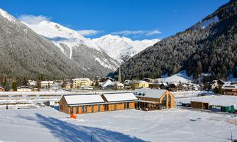 A snowy landscape with mountains and a clear blue sky. In the foreground, there is a modern wooden building in a peaceful setting.