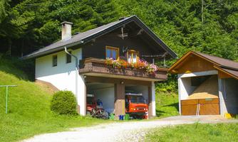 A cozy house in nature with a balcony full of flowers. Next to it is a barn, surrounded by green meadows and trees.