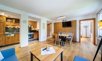 A modern living room with an open kitchen and wooden furniture. The space is bright and inviting, with a dining table and comfortable sofas.