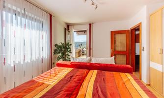 A cozy bedroom with colorful bed covers and large windows. A plant and a view of the scenery can also be seen.