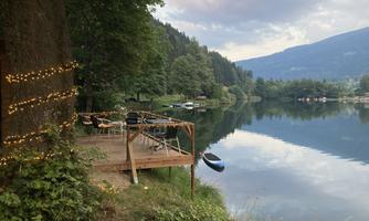 A calm lake with a wooden terrace and chairs. Surrounded by trees and gentle hills, the scene exudes a relaxed atmosphere.