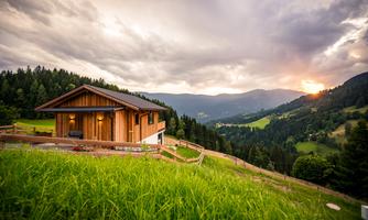 A modern wooden house in a landscape with gentle hills. The sun sets behind the mountains and the sky is cloudy.
