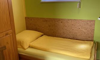 A simple room with a single bed and a yellow blanket. The walls are painted green, and a wooden wardrobe is visible in the background.