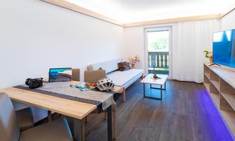 A modern living room with a sofa, a table, and a television. The room has large windows that look out onto a balcony and is bright and inviting.