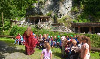 Eine Gruppe von Kindern sitzt im Freien und schaut aufmerksam einer Erzählerin in einem roten Gewand zu. Im Hintergrund sind Felsen und Holzstrukturen sichtbar.