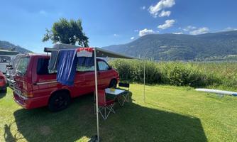 A red car is standing on a green meadow with a sun umbrella. In the background, gentle hills and a blue sky can be seen.