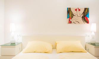 A modern bedroom with a large bed and yellow pillows. A colorful artwork of a woman making a "shh" sign with her finger on her lips hangs on the wall.