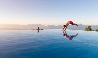 A beautiful infinity pool with a view of the mountains. One person jumps into the water while another relaxes swimming.