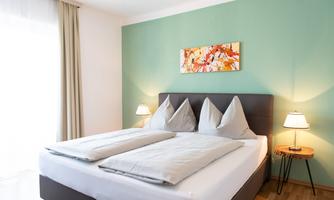 A cozy bedroom with a large bed and green wall color. There are bedside lamps and a decorative picture on the wall.