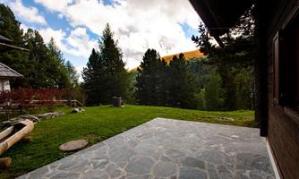 A tranquil landscape with green grass and tall trees. In the background, mountains and a blue sky with clouds can be seen.