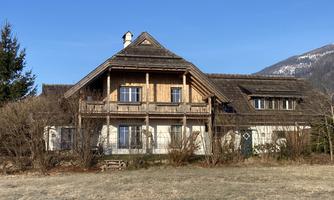 A charming country-style house with a wooden balustrade. Surrounded by a meadow and mountains in the background.