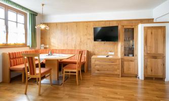 A cozy dining area with wooden furniture and a bench. There is a television on the wall and large windows provide daylight.