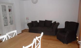 A cozy living room with a dark brown sofa and an armchair. There is a wooden table with white chairs and light walls.