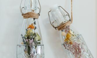 Two decorative glass bottles with dried flowers, hanging on a wall. The bottles are adorned with a jute ribbon.