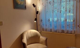 A bright room with a cozy chair and a window with curtains. There is a picture hanging on the wall, and there is a lamp next to the chair.