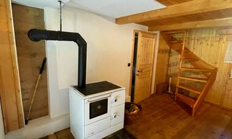 A cozy room with a wood stove and wooden floor. In the background, a spiral staircase can be seen.