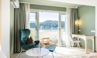 A modern room with a view of the mountains and a balcony. There is a comfortable armchair, a table, and plenty of natural light.