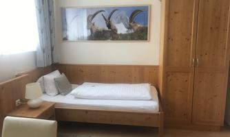 A simple bedroom with a single bed and a wooden dresser. On the wall hangs a picture of ibexes in an alpine landscape.