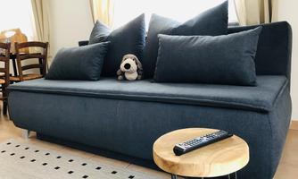 A cozy, dark blue sofa with several pillows and a small stuffed animal. Next to it is a round table with a remote control.