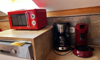 A compact kitchen corner with a red microwave, a coffee machine, and a kettle. The appliances are placed on a light wood shelf.