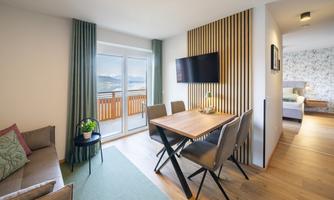 A modern living room with a dining table and chairs. Large windows offer a view of the scenery outside.