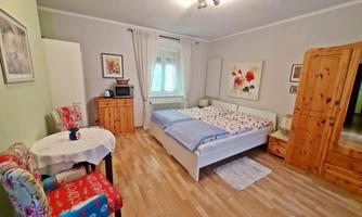 A cozy bedroom with a double bed, a table, and a chair. The walls are light and there are decorative pictures.