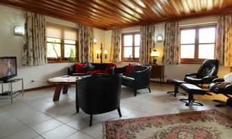 A cozy living room with wooden floors and white walls. There are comfortable furniture, large windows, and a television.