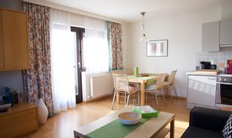 A bright and cozy living space with a dining table and chairs. The windows are adorned with curtains and the floor is made of wood.
