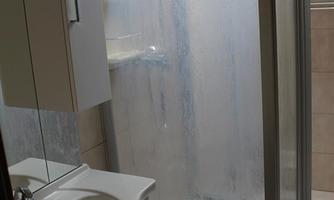 A bathroom with a glass shower and a sink. The walls are tiled and there is daylight coming through a window.