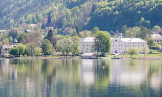 A quiet lake with clear reflections of the surrounding landscape. In the background, historical buildings and a church are nestled among green trees.