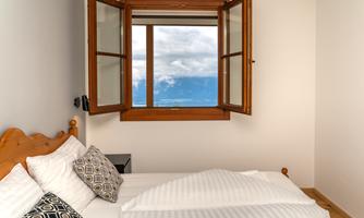 A cozy bedroom with a window that offers a view of cloudy mountains. The bed is decorated with white and patterned pillows.