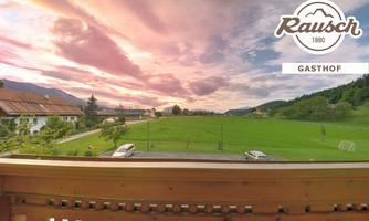 A picturesque view featuring a meadow and gentle hills under a colorful sunset. In the foreground, there is a balcony with a wooden railing.