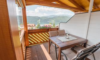 A balcony with a wooden table and chairs. The view extends over a beautiful landscape with mountains and a clear sky.