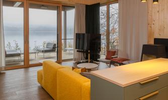 A modern living room with a yellow sofa and a large window. In the background, there is a tranquil lake and a terrace.