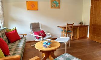 A cozy living space with a sofa, an armchair, and a table. In the background, there are decorative pictures on the wall and a wooden shelf.