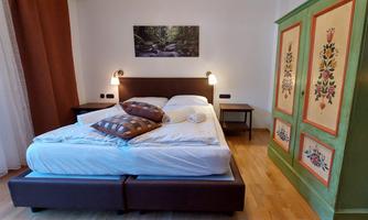 A cozy bedroom with a large bed and colorful pillows. In the background, there is an artistically painted wardrobe and a picture of a natural landscape.