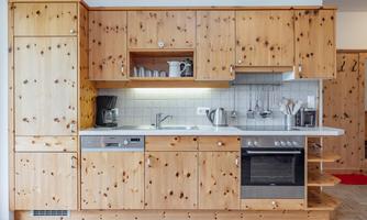 A modern kitchen made of light wood with white tiles. It is equipped with a refrigerator, stove, and sink.