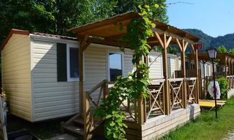 A charming holiday home with a wooden porch, surrounded by green trees. The surroundings are calm and inviting.