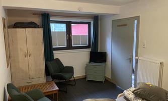 A simple room with a bed, a chair, and a small table.  
There is a wardrobe against the wall and a television is available.