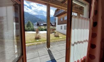A view from a window onto a well-maintained courtyard with paved paths and green grass. In the background, mountains and houses can be seen.