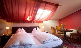 A cozy bedroom with a large bed and red-transparent curtains. In the background, there is a table with chairs and a decorative plant.