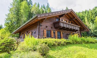 A cozy wooden house surrounded by green trees and bushes. The sky is slightly cloudy and gives a calm impression.