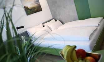 A modern bedroom with a double bed and fresh white bed linens. In the foreground, there are fruits and plants that create an inviting atmosphere.