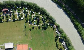 An aerial image of a recreational area by the river. Surrounding the green meadows are parking spaces for motorhomes and trees.