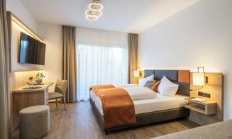 A modern hotel room with two beds, a desk, and a television.  
Large windows provide plenty of daylight and a bright atmosphere.