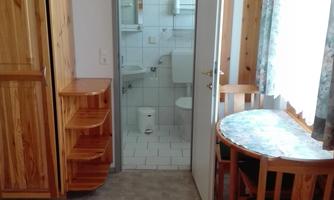 A simple room with wooden furniture and a table with chairs. In the background, a bathroom is visible.